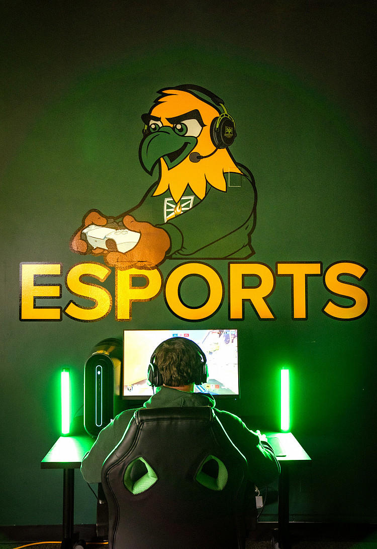 Student with a gaming headset on, sitting in a gaming chair, and playing video games in our on-campus eSports room.