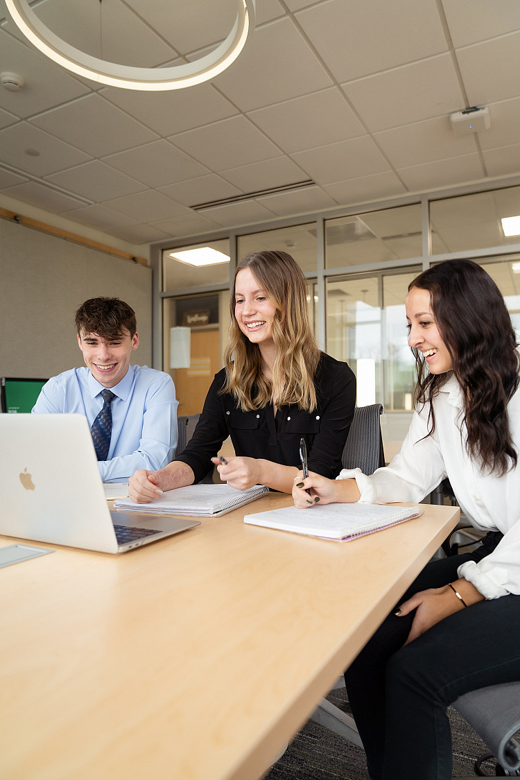 Three students sitting at a table with business casual clothing on and looking at a laptop