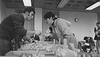 Two people leaning over a table toward each other, handing something between them.