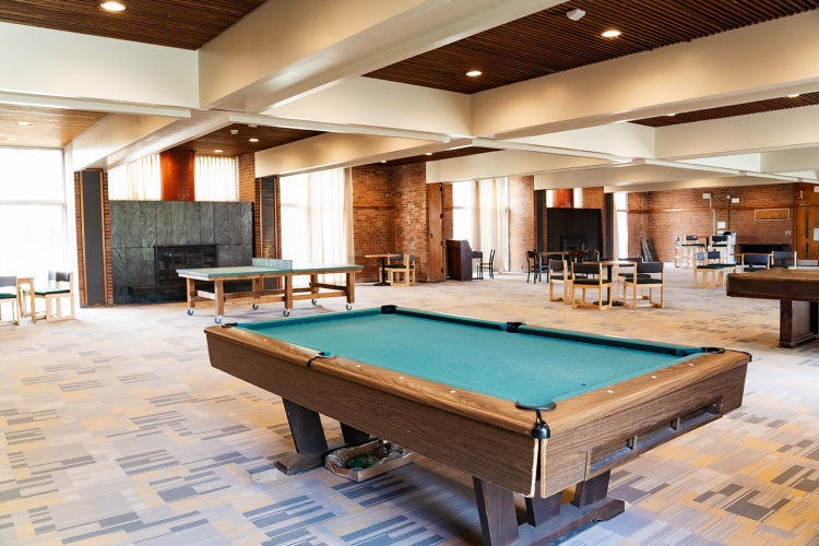 Close up of a pool table with a ping pong tables, chairs and smaller tables in the background of a common room