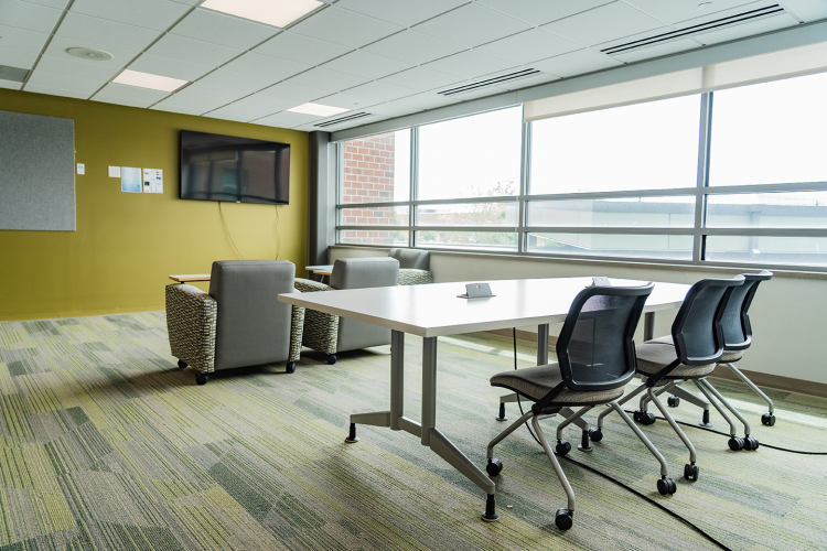 Angled view of a lounge area with armchairs and a table with office chairs, facing a wall-mounted TV