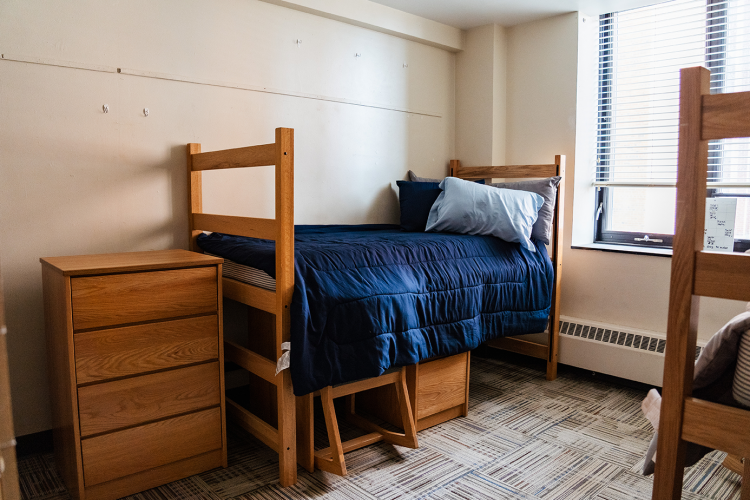 Angled view of a bedroom with a dresser, bed, and a desk with chair underneath the bed